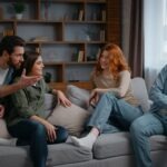 Multiracial Four Friends At Home Party Talking On Couch Emotional Discussion Arguing Smiling Diverse People Communicate In Living Room Women Girls Men Guys Together Conversation Debate Problem Discuss